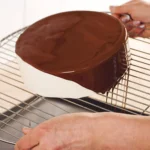 Chocolate glaze being poured over a round cake on a stainless steel cooling grid
